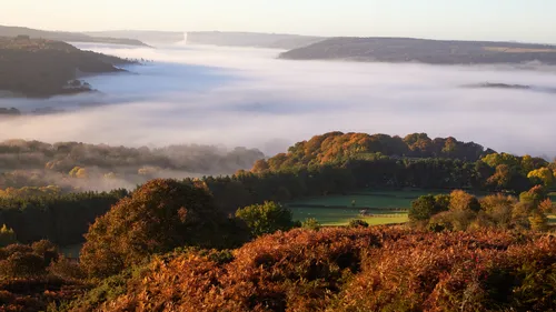 This landscape photograph captures the natural beauty of the Peak District in Derbyshire, United Kingdom, during an autumn morning at sunrise. The image shows a rural scene with Curbar Edge in the foreground, where bracken and trees display vibrant autumn colours. A dramatic cloud-inversion effect is visible, with mist and low-lying clouds blanketing the valleys and separating the wooded hills. The photograph highlights the unique character of nature within the Peak District, as the rising sun casts warm light across the landscape. This morning scene emphasizes the atmospheric conditions commonly found in autumn, with mist creating a tranquil and picturesque moment in the countryside.